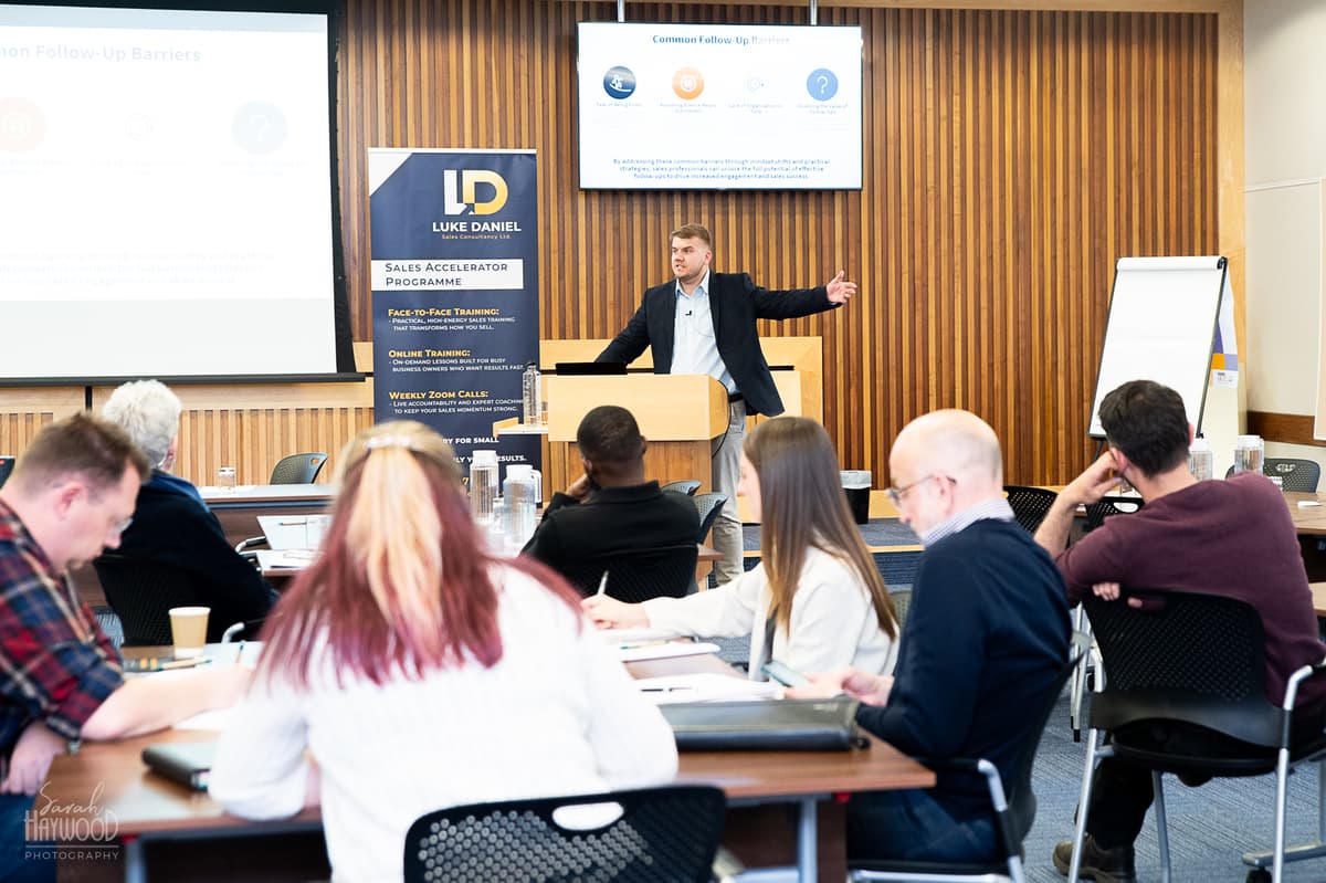 Luke speaking at a podium while attendees sit around tables at a training session
